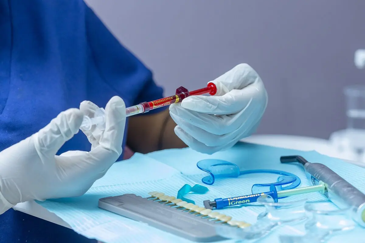 Gloved hands holding a gel syringe over a tray holding a lip retractor, a shade guide and clear trays